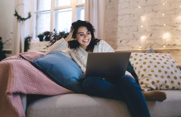 圣诞节期间 穿着保暖毛衣和牛仔裤的乐观女性坐在舒适的沙发上 坐在垫子里 一边笑着一边用笔记本电脑 — 图库照片
