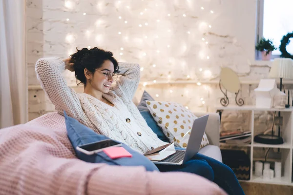 Side view of positive young female in warm outfit and eyewear sitting on cozy sofa with laptop and notebook on knees