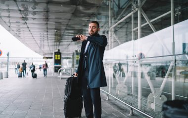 Bavul taşıyan sakallı yakışıklı işadamı modern binanın yanında duruyor ve yolculuk sırasında araç bekleyen saate bakıyor.
