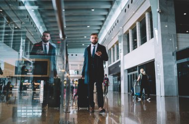 Havaalanındaki terminal kapısını ararken kameraya bakan bavul ve akıllı telefon teknolojisiyle şirket yöneticisinin tam boy portresi, iş gezisi sırasında Kafkasyalı işveren 30 'lu yaşlarda.