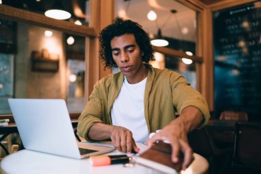 Düşünceli kıvırcık erkek öğrenci sınavlara hazırlanıyor ve kafeteryada teknolojiyle ödev yapıyor. Düşünceli adam serbest çalışıyor. Bilgisayarda uzaktan çalışıyor ve veri tarıyor.