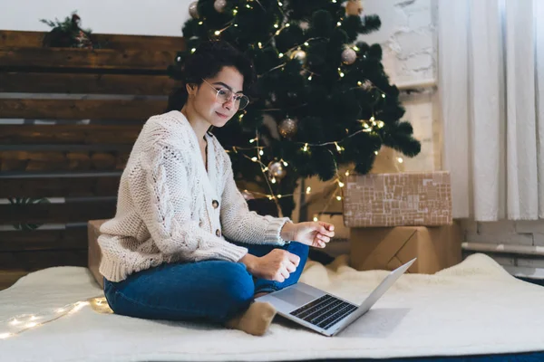 Full body of young lady in warm wear sitting with legs crossed on floor under Christmas tree and surfing laptop