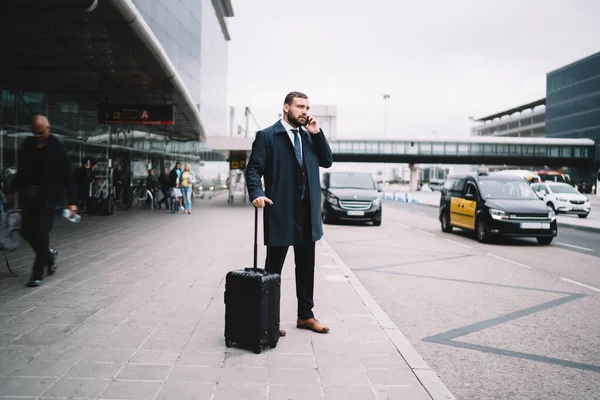Caucasian businessman using mobile phone for calling after arrival to airport terminal, male lawyer with baggager using cellphone application for contact customer service and order cab taxi