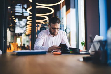 Optik göz giyimli genç serbest çalışan masa başına oturmuş uzaktan kumandalı iş sırasında akıllı telefon tebligatı kuruyor, genç bir erkek cep telefonuyla ilgili bilgi arıyor