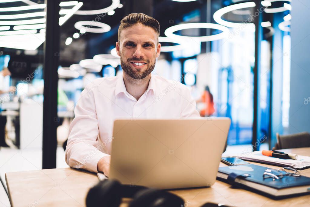 Portrait of Caucasian male IT developer installing app on laptop computer sitting at table desktop and smiling at camera, skilled freelancer using netbook for web remote job in coworking space
