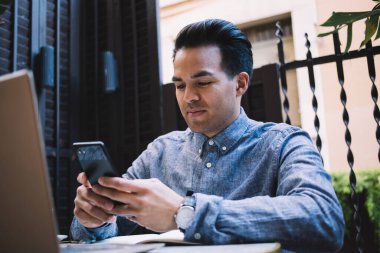 Düşünceli, serbest çalışan, serbest çalışan bir etnik erkek, laptopla masada oturuyor ve açık kafedeki akıllı telefondan mesajlaşıyor.