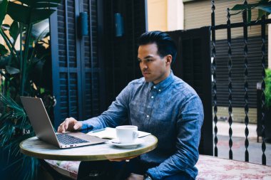Düşünceli etnik erkek serbest çalışan günlük kıyafetlerle açık kafedeki yuvarlak masada oturuyor ve dizüstü bilgisayarla uzaktan kumandalı bir proje üzerinde çalışıyor.
