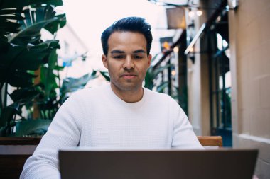 Ciddi etnik erkek serbest çalışan, günlük kıyafetleriyle açık kafede oturuyor ve gündüzleri bilgisayarını karıştırıyor.