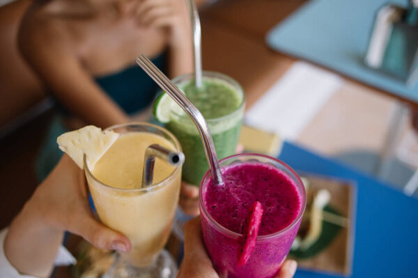 High angle of crop faceless group of female friends relaxing in cafe and cheering glasses of fresh healthy smoothies served with dragon fruit lime and pineapple slices