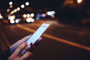 Bilinmeyen bir kadının cep telefonunun yan görüntüsü. Gece şehrinde arabaların bulanık ışıklarının yanında dururken.