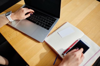 Yukarıda akıllı saati olan, dizüstü bilgisayarda yazan ve ahşap masada elinde kalem ve defterle oturan isimsiz bir kadından.