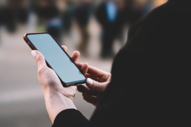 Siyah ceketli tanınmayan bir kadının şehirde dikilirken cep telefonuna bakması ve yoldan geçen insanların siluetleri.