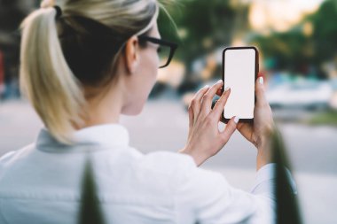 Kısa saçlı, beyaz tişörtlü ve gözlüklü, güneşli bir günde sokakta dikilirken cep telefonuyla fotoğraf çeken mahsul kadınların arka planı.