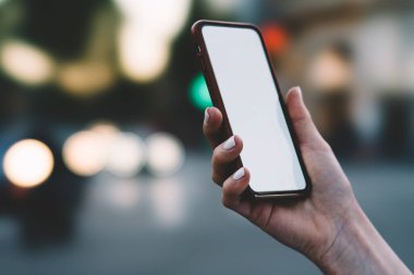 Bulanık sokak arka planında cihazı elinde tutarken boş ekranlı modern kablosuz cep telefonu kullanan kimliği belirsiz bir kadın.