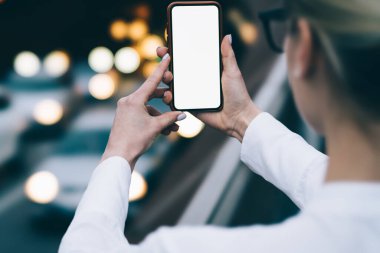 Resmî beyaz gömlek ve gözlük takan yüzü olmayan bir kadının aydınlanmış caddede dikilip cep telefonuyla fotoğraf çekişi.