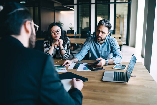 Male manager pointing at document and asking for opinion of colleague while having meeting of multiracial business team in office