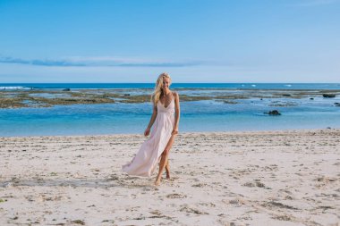 Güzel, gülümseyen, yalın ayaklı, uzun boyunlu, beyaz elbiseli, kumlu sahilde yürüyen bacaklarını gösteren sarışın bir kadın.