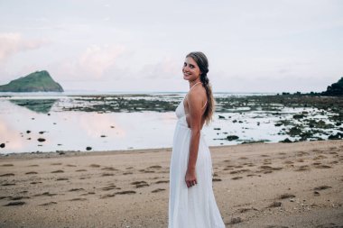 Güneşli bir günde kumlu sahilde gülümseyen ve gezinen beyaz elbiseli, uzun saçlı neşeli sarışın kadının yan görüntüsü.