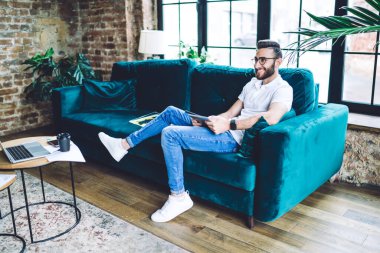 Yan görünüşte mutlu bir erkek rahat mavi koltukta rahatlar ve tablet kullanır. Bu arada başka yere bakar ve yaratıcı bir dairede dinlenir.