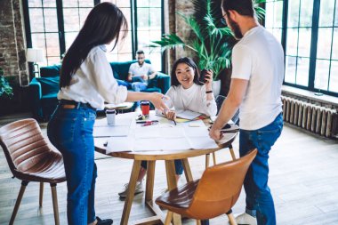Beyaz tişörtlü, çok ırklı meslektaşlar gazetelerle çalışırken iş stratejisi hakkında konuşuyor ve ofiste kahve içmenin keyfini çıkarıyor.