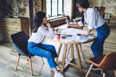 Düşünceli çok ırklı kadınlar gündelik giysiler içinde ahşap yuvarlak masada oturuyor ve modern çatı katında çalışmak için toplanıyorlar.