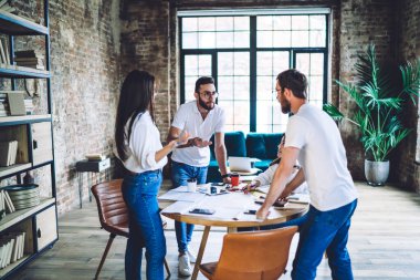 Bir grup meslektaş, büyük pencereli modern projenin yaratıcı fikirleri üzerinde kafa patlatırken ayakta ve oturuyorlar.