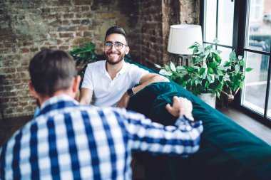 Moden Ofis 'te parlak bir günde pencerenin yanında oturmuş iş arkadaşıyla sohbet eden beyaz tişörtlü, gülümseyen bir yönetici.