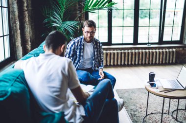 Sakallı, odaklanmış, rahat giyinen ve gözlüklü erkek iş arkadaşları kanepeye oturup proje üzerinde birlikte çalışıyorlar.