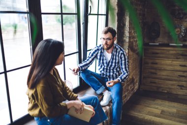 Ciddi çoklu etnik gruptan iş arkadaşları günlük kıyafetlerini giyip yaratıcı işyerlerinde pencere eşiğine oturmuşlar ve birbirleriyle konuşuyorlar.