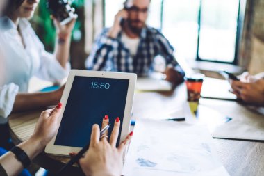 Tablet ekranın yumuşak odağı, kafede masada oturan ve iş stratejisini iş arkadaşlarıyla tartışan bir kadının ellerinde.