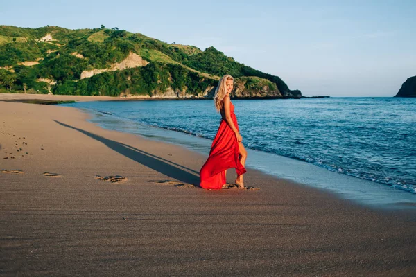 Mavi deniz ve yeşil tepe boyunca sakin kumlu sahilde yürürken parlak kırmızı elbiseli neşeli kadının yan görünüşü bacağını gösteriyor.