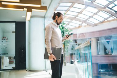 Neşeli beyaz erkek çalışan cep telefonuyla haberleri okuyor ve kontrol ediyor modern ofis içi, başarılı genç girişimci web sitesi e-posta gönderiyor.