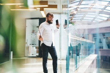 Ofisteki çalışma yerinde ayakta duran, gülümseyen, başarılı, sakallı, şık giyinmiş, kapalı mekandan telefon eden neşeli yönetici.