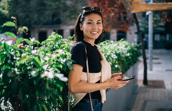 Half length portrait of asian female blogger using modern mobile phone for communication outdoors, smiling teen woman millennial standing outdoors holding smartphone connected to 4G internet