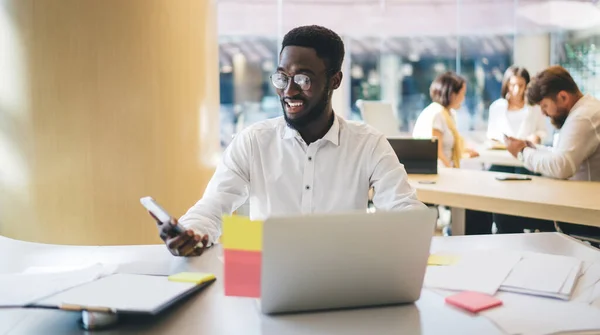 Happy dark skinned male in glasses for vision protection checking mobility during remote job on laptop technology in modern coworking space, cheerful man enjoying smartphone messaging indoors