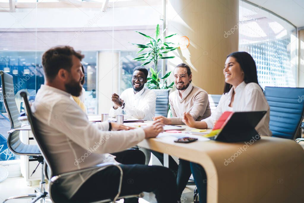 Group of multiethnic delighted coworkers in formal wear sitting at table in creative workspace and discussing startup ideas while laughing and looking at each other