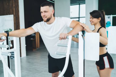 Beyaz spor tişörtlü ve siyah şortlu genç adam spor simülatöründe egzersiz yapıyor ve kadın eğitmen pozisyonunu kontrol ediyor.