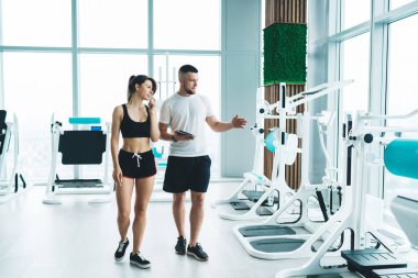 Sakallı kişisel antrenör elinde tabletle spor kıyafetli genç bir bayanla yürüyor ve modern fitness stüdyosundaki spor aletlerini anlatıyor.