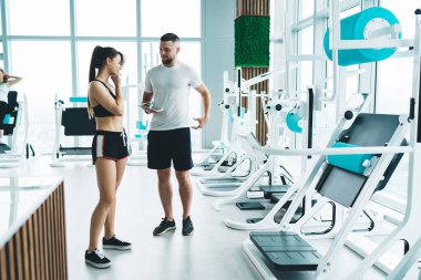 Siyah spor tişörtlü ve şortlu gülümseyen genç sporcunun yan görünüşü ve geniş spor salonunda erkek kişisel koçu dinlemesi.