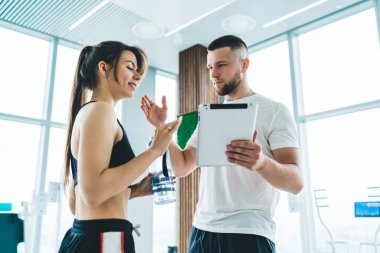 Modern hafif spor salonunda durup gülümseyen bir sporcuyla konuşan ciddi sakallı spor ayakkabısı ve tablet eğitmeni.