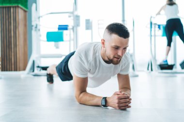 Akıllı saat odaklı sporcu gündüz vakti yoğun ısınma sırasında modern spor salonunun ortasında dirsek tahtası yapıyor.