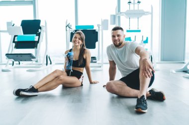 Sağlam vücutlu genç bir erkek ve kadın yerde dinleniyor ve yoğun eğitimden sonra spor salonunda kameraya gülümsüyor.