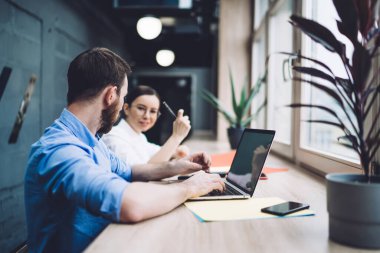 Erkek çalışanın dizüstü bilgisayarına göz atması ve masa başında oturup birlikte proje üzerinde çalışırken kadın iş arkadaşını dinlemesi.