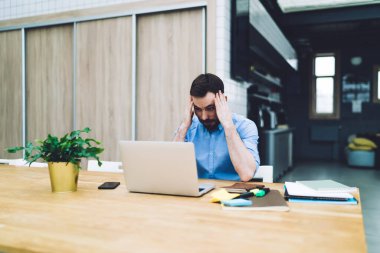 Üzgün sakallı iş adamı, iş yerinde masada otururken ellerini başının üstüne koyup dizüstü bilgisayara bakıyor.