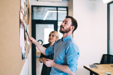 Modern iş yerinde duvarda grafik gösteren kendinden emin genç erkek işçinin yan görünüşü kalemle kadın yöneticiye.