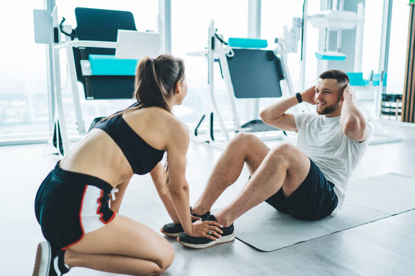 Female trainer helping man in doing torso lifting while holding his feet during training in modern gym with panoramic windows