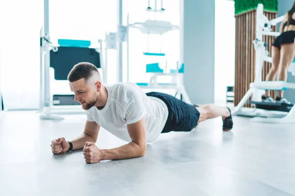 現代の軽体育館でワークアウトをしながら 床の上に板運動を行うアクティブウェアの強力なひげそりのスポーツマンの完全な体 — ストック写真