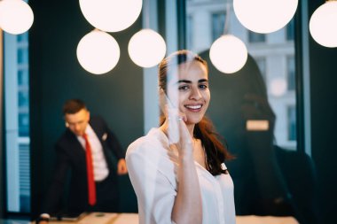 Ofisteki cam duvarın yanında dururken kameraya bakan ve cep telefonuyla konuşan genç bir kadın çalışanın penceresinden.