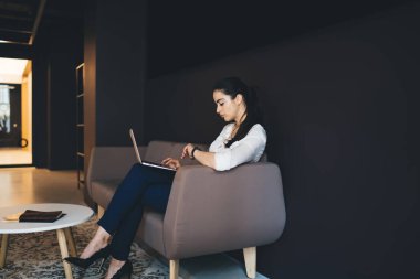 Proje sırasında rahat koltukta oturan ve dizüstü bilgisayarları karıştıran resmi kıyafetli konsantre etnik kadın yöneticinin yan görüntüsü