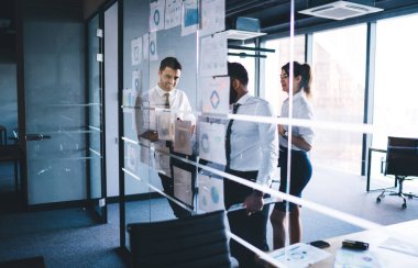 Başarılı erkek ve kadın ortaklar, kağıt gelir raporlarıyla cam standının yanında duran sermayenin parasal kazancını analiz ediyor ve takas deneyimi, işbirliği süreci için görüşmeler yapıyor.